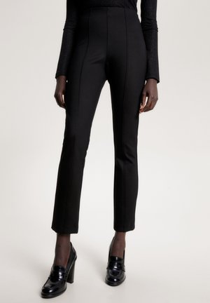 Black high-waisted trousers with a slim fit, smooth fabric, vertical seams, and ankle-length cut, paired with black heeled loafers.