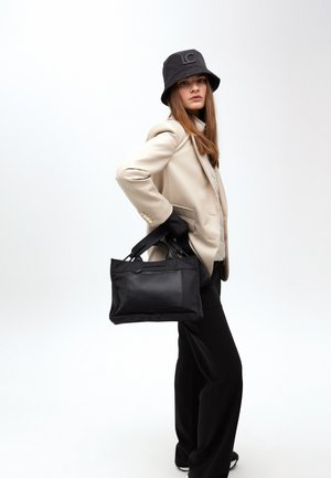 Modell in einem beigen Blazer und schwarzem Bucket Hat hält eine schwarze Handtasche und posiert selbstbewusst vor einem neutralen Hintergrund.