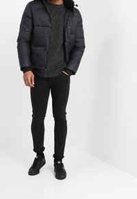 Černá péřová bunda s kapucí, texturovaný šedý svetr a přiléhavé černé džíny. Černé tenisky s bílými akcenty dotvářejí celkový outfit.