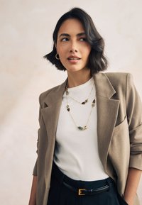 Layered gold necklaces with teal accents, worn over a ribbed white shirt and beige blazer, paired with black high-waisted trousers.