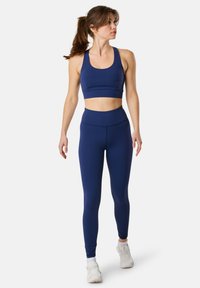 Sportivo color blu navy con reggiseno e leggings a vita alta realizzati in tessuto elastico. Presentano un design aderente e una texture liscia. Scarpe da ginnastica bianche.