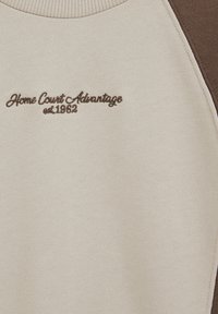 Sveter s farebnou kombináciou krémovej a hnedej. Má rebrovaný výstrih a hnedý vyšívaný nápis "Home Court Advantage est. 1962".