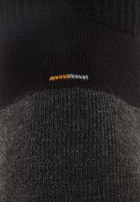 Calcetín negro hasta el tobillo con textura acanalada y talón reforzado en contraste. Pequeño detalle de costura naranja y azul cerca de la parte superior.
