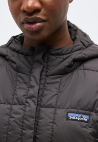 Primo piano di una persona che indossa una giacca trapuntata nera Patagonia con cappuccio, con il logo del marchio sul petto.