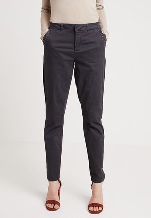 Femme portant un pantalon noir moulant avec des poches cloutées et un haut beige à manches longues, assortis à des talons ouverts marron, debout droit.