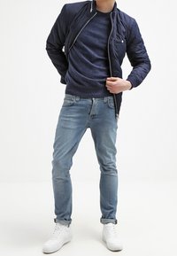 Osoba ubrana w granatową pikowaną kurtkę na ciemnoniebieskim swetrze, jasnoniebieskie podwinięte dżinsy i białe trampki, stojąca na tle białego tła.