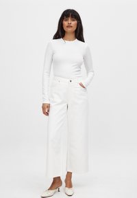 Witte langemouwtop gecombineerd met brede witte broek. De top heeft een geribbelde structuur; de broek heeft een klassieke vijf-pocketdesign en is onversierd.