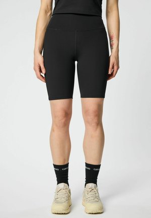 Frau trägt schwarze Biker-Shorts mit hoher Taille, schwarze Socken mit dem Schriftzug „GIRLFRIEND“ und beige Sportschuhe, steht vor einem einfarbigen Hintergrund.