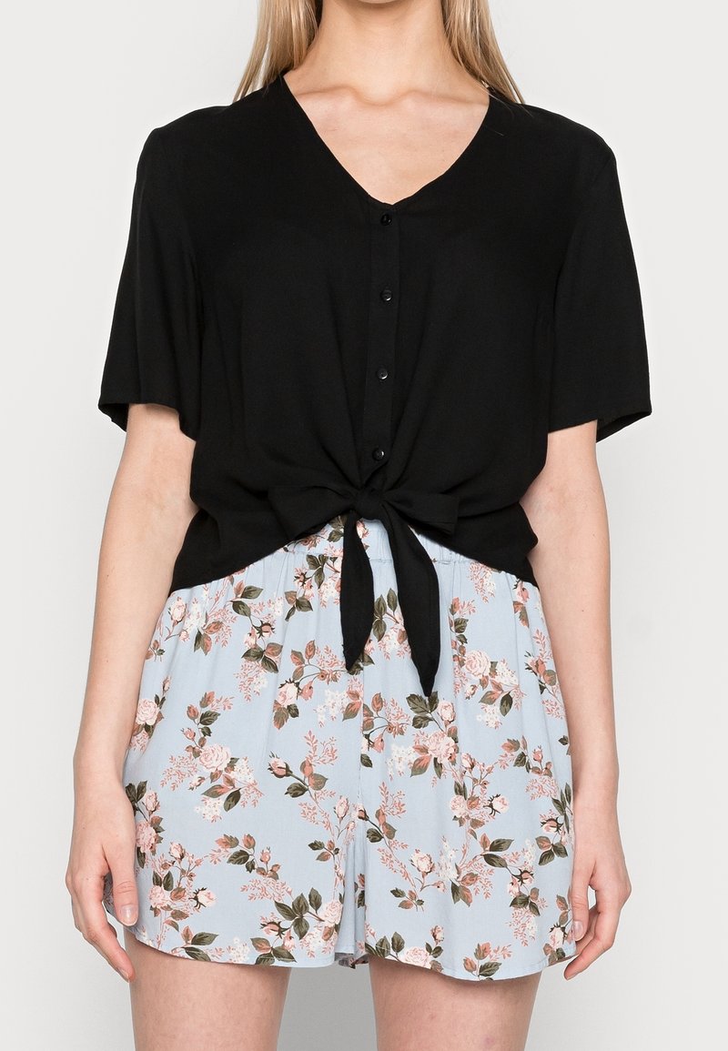 Femme portant un haut noir à manches courtes boutonné, noué à la taille, et un short bleu clair à motif floral.