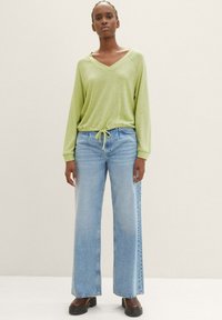 Grünes Langarm-V-Ausschnitt-Shirt mit Kordelzug, kombiniert mit hellblauen, weiten Jeans und schwarzen Plateaustiefeln. Glatte Textur, lässiges Design.