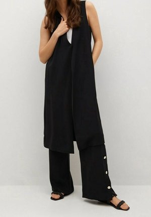 Vrouw draagt een lange zwarte mouwloze vest over wijde zwarte broek met witte zijknopen en zwarte gevlochten sandalen tegen een effen achtergrond.