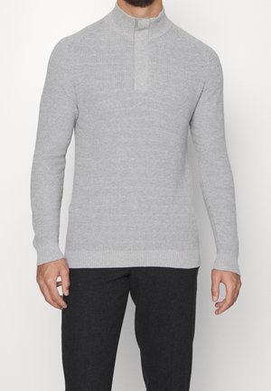 Homme portant un pull-over en maille côtelée gris clair avec col montant et pantalon noir, debout les bras détendus.