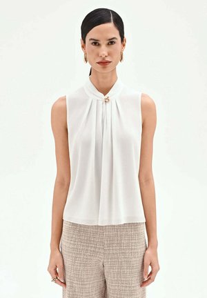 Donna con capelli scuri raccolti in una coda di cavallo liscia, che indossa orecchini a cerchio dorati, una camicetta bianca senza maniche con una chiusura a perla e pantaloni beige texturizzati.