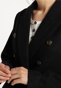 Czarny, dwurzędowy wełniany płaszcz z metalowymi guzikami, wykonany z teksturowanej tkaniny, z szerokimi klapami, oraz zbliżenie na ręce regulujące dopasowanie.