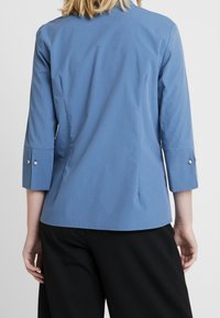Lichtblauwe blouse met driekwartmouwen, voorzien van knoopmanchetten en een eenvoudig, gestroomlijnd ontwerp. Gemaakt van een zachte stof.