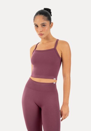 Junge Frau, die in einem bordeauxfarbenen enganliegenden Croptop und hoch taillierten Leggings mit zu einem Dutt gebundenen Haaren vor weißem Hintergrund steht.