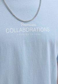 Svijetloplavi pamučni majica s natpisom "Premium COLLABORATIONS" u bijeloj boji, s vidljivim srebrnim lančićem iznad.