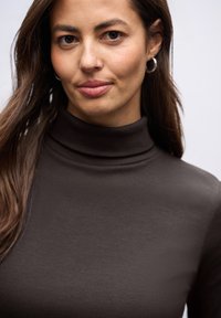 Czarny prążkowany golf, z wysokim kołnierzem i gładką fakturą. Modelka ma długie włosy i srebrne kolczyki w kształcie kół. Miękkie, jasne tło.