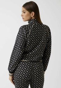 Zwarte trainingsjas met een overal goudkleurig geometrisch patroon, een hoge kraag en geribbelde manchetten met gestreepte accenten.