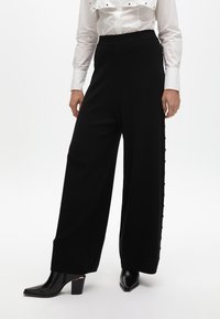 Černé kalhoty s širokými nohavicemi a hladkým povrchem, zdobené zlatými knoflíky po stranách, v kombinaci s bílou košilí s límečkem a černými lodičkami.