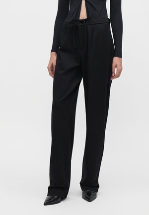 Vrouw draagt zwarte wijde broek met hoge taille en een gebonden riem, gecombineerd met zwarte puntige schoenen en een zwarte top met lange mouwen.