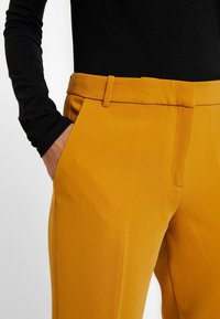 Spodnie w kolorze musztardowym, dopasowane, o gładkiej fakturze, z umiarkowanie wysokim stanem, paskami na pasek i bocznymi kieszeniami, zestawione z czarną bluzką z długim rękawem.