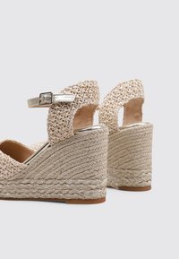 Sandalias de cuña trenzadas color beige con talón abierto, correa al tobillo asegurada por una hebilla plateada y una suela de cuerda texturizada.