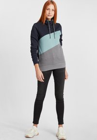 Le sweat à capuche présente un design en blocs de couleurs marine, bleu clair et gris, avec une coupe décontractée, un cordon de serrage et des poignets et un ourlet côtelés.