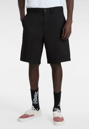 Mand iført sorte knælange shorts, sorte sokker med hvid tegneserie-kanin, og hvide og lyserøde sneakers, stående mod en hvid baggrund.
