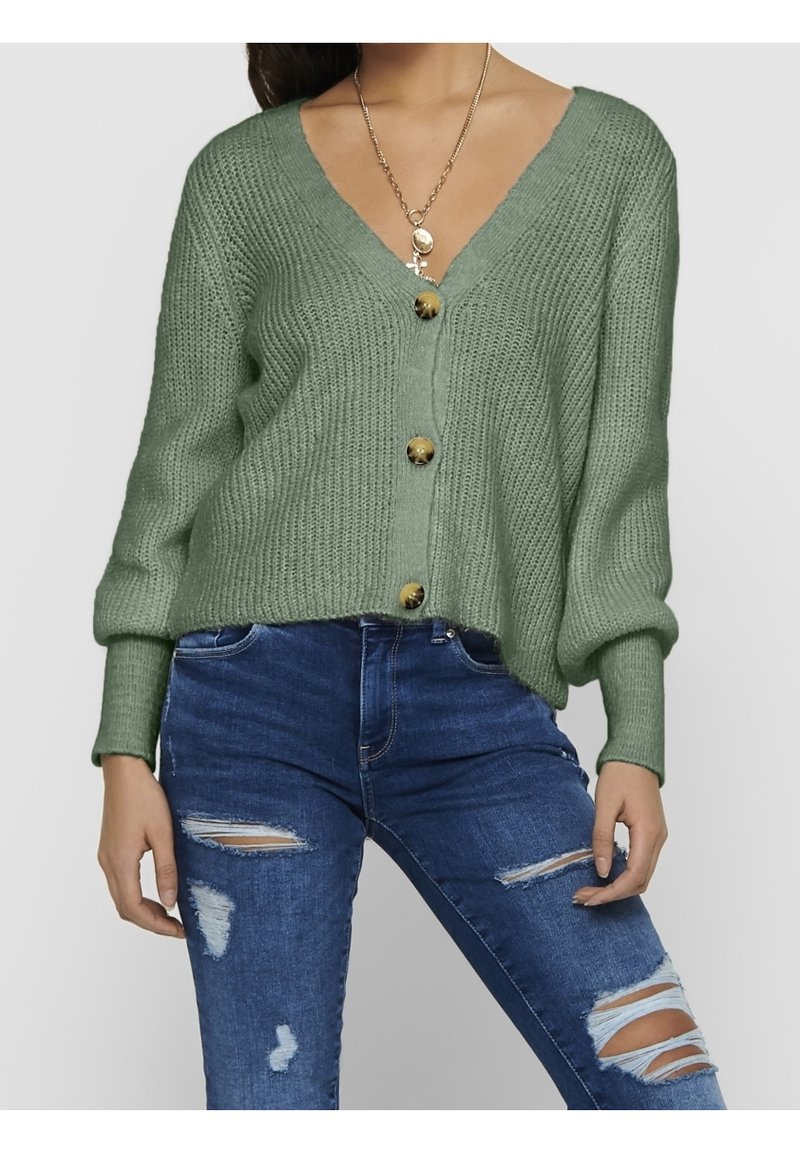 Femme portant un cardigan tricoté vert clair boutonné, un collier en or et un jean skinny bleu déchiré sur un fond uni.
