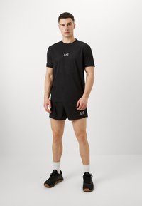 Μαύρο αθλητικό μπλουζάκι με υφή και λογότυπο EA7, συνδυασμένο με μαύρη βερμούδα και μαύρα αθλητικά παπούτσια, με λευκές κάλτσες.