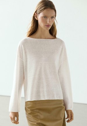 Donna che indossa un maglione rosa chiaro a maniche lunghe e una gonna di pelle marrone, in piedi davanti a uno sfondo chiaro e uniforme.