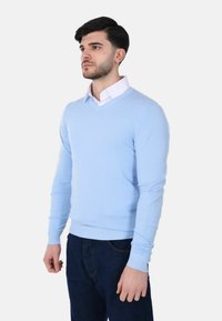 Uomo con capelli corti e scuri che indossa un maglione azzurro chiaro a V sopra una camicia bianca con colletto e jeans blu scuro, su uno sfondo neutro.