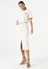 T-shirt en coton beige avec graphique rouge ; jupe en denim blanche avec fente avant ; sandales compensées beiges. Design simple et décontracté avec une coupe relaxante.