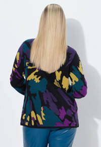Blonde Frau mit glattem Haar trägt einen schwarzen Pullover mit türkisfarbenen, lila und gelben abstrakten Mustern sowie eine blaue Lederhose und steht mit dem Rücken zur Kamera.