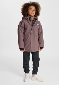 Giacca impermeabile viola con cappuccio, cerniera frontale e due grandi tasche. Abbinata a pantaloni jogger neri e sneakers bianche.