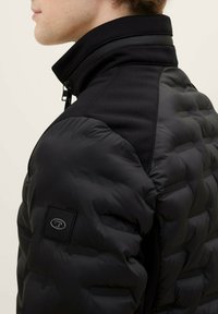 Giacca imbottita nera con colletto alto, pannelli texture sulle spalle e un'etichetta con logo sul braccio sinistro. Tessuto liscio e elegante.