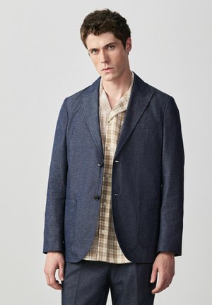 Muž v tmavomodrom saku cez béžovú kockovanú košeľu, stojaci pred jednoduchým svetlosivým pozadím.