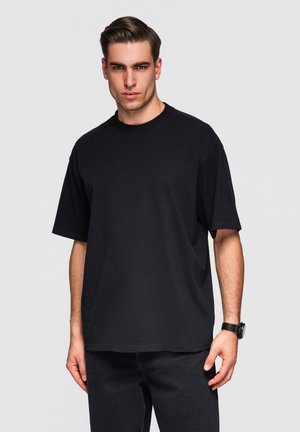 Jeune homme aux cheveux courts et foncés portant un t-shirt noir uni oversize et un pantalon noir sur un fond gris clair.