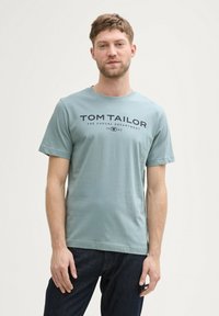 Šviesiai mėlyna medvilninė marškinėlių su trumpomis rankovėmis ir apvaliu iškirpimu. Priekyje yra tamsiai mėlynos spalvos atspausdintas tekstas "TOM TAILOR".