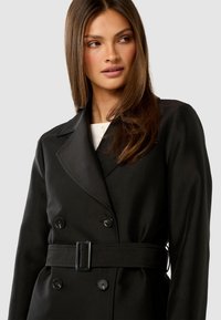 Woman with long brown hair wearing a black belted double-breasted coat and white top, looking to the side against a plain background.
