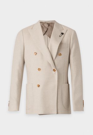 Young man standing wearing beige double-breasted blazer with gold buttons, white shirt, dark brown trousers, and dark brown shoes.