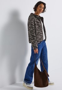 Femme se tenant à l'intérieur, vêtue d'une veste en polaire à motifs, d'une chemise blanche, d'un jean bleu et de baskets blanches, tenant un grand sac à bandoulière en daim marron.