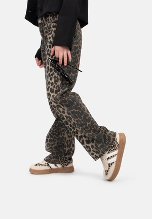 Pantalons en denim imprimé léopard beige et noir, associés à des baskets beiges avec des accents et des rayures noires, tenant des lunettes de soleil à la main.