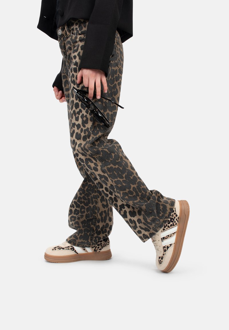 Pantaloni in denim con stampa leopardo beige e nera, abbinati a sneaker beige con accenti e strisce nere, reggevano occhiali da sole in mano.