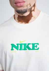 Cremefarvet t-shirt med et stort grønt og neongult "NIKE" logo og teksten "NURTURE THE SOLE" trykt på forsiden.