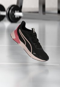 Μαύρο και ροζ παπούτσι running Puma με λευκή σόλα, αιωρείται πάνω από έναν φωτεινό γυμναστηριακό δάπεδο με θολό βαράκι στο φόντο.