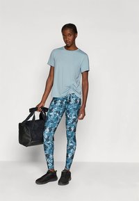Lichtblauw T-shirt met korte mouwen en een knoopdetail. Donker gebloemde legging in blauwtinten, gecombineerd met zwarte sportschoenen en een sporttas.