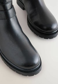 Botas de cuero negro hasta el tobillo con una textura suave, punta redondeada y suelas de goma gruesa. Cuenta con una correa decorativa alrededor del tobillo.