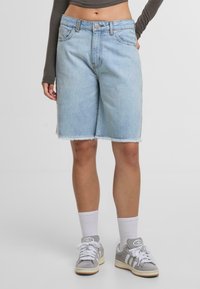 Persona que lleva pantalones cortos de mezclilla azul claro hasta la rodilla con dobladillos deshilachados, calcetines blancos, zapatillas grises de Adidas y una camiseta de manga larga gris.
